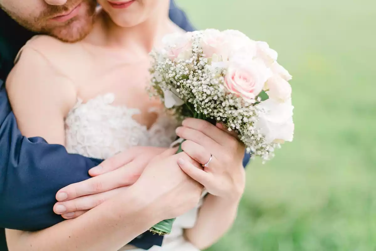 Una mujer vestida de boda sostiene un ramo de flores abrazada a su marido